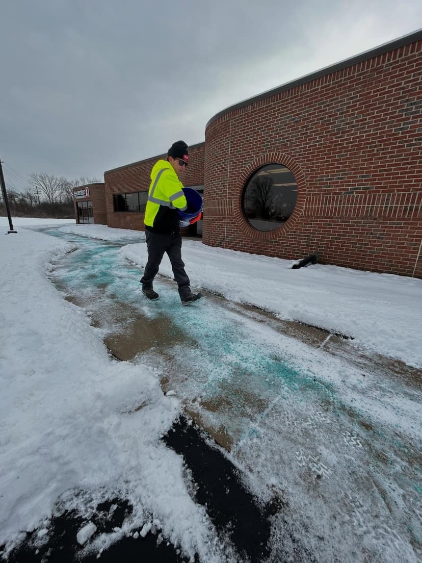 Worker spreading salt on snowy sidewalk near brick building to prevent ice accumulation.